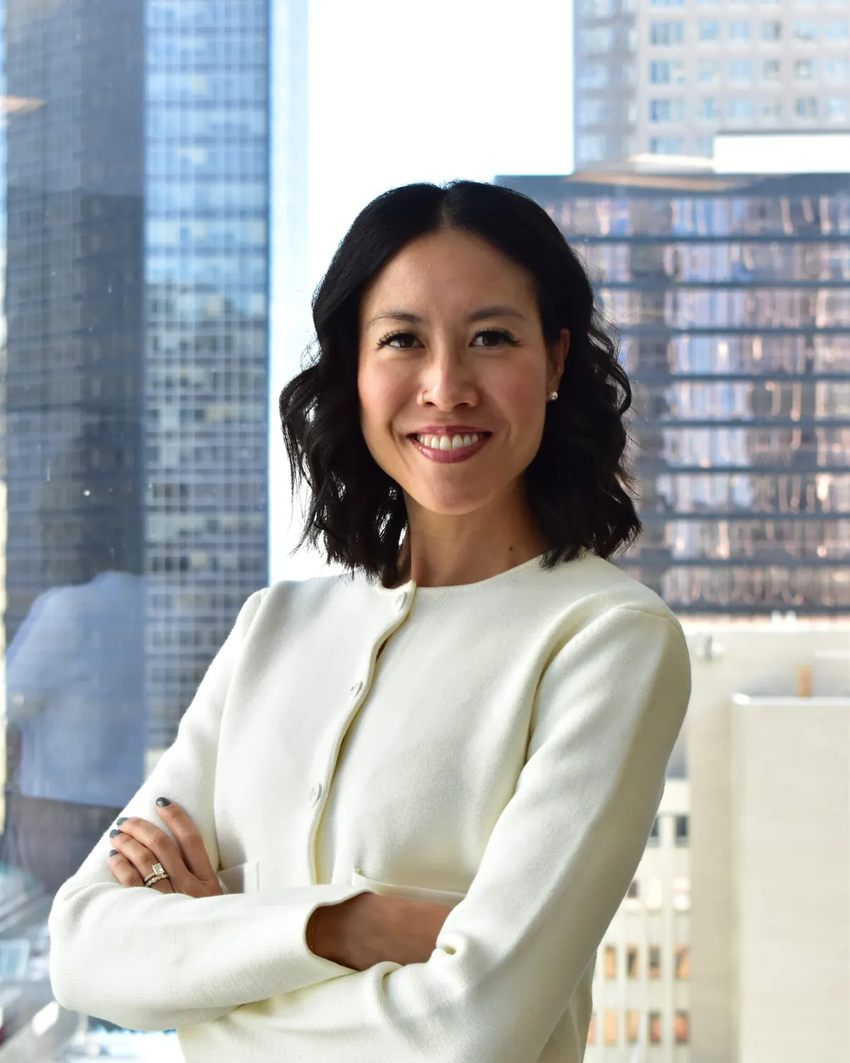 Denise Yee in a cream jacket, arms folded, smiling in front of a downtown Calgary office window