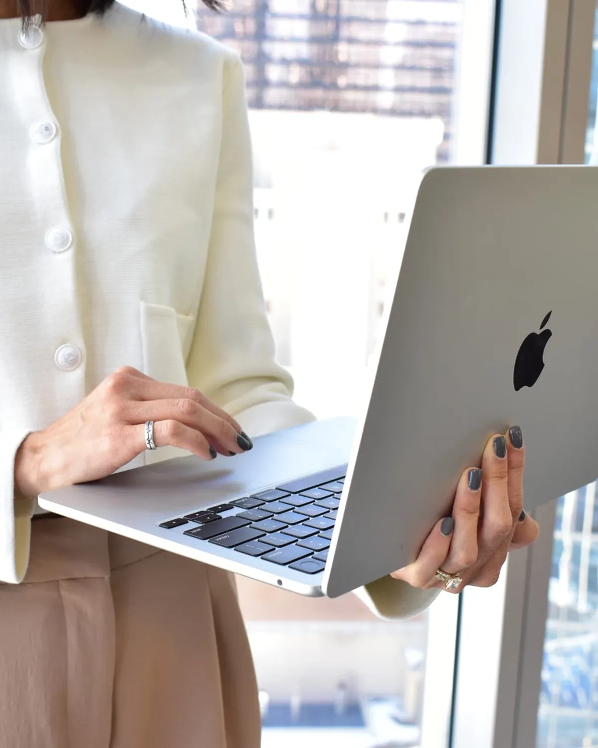 Denise holds an open laptop while standing at a downtown Calgary office window, typing with one hand