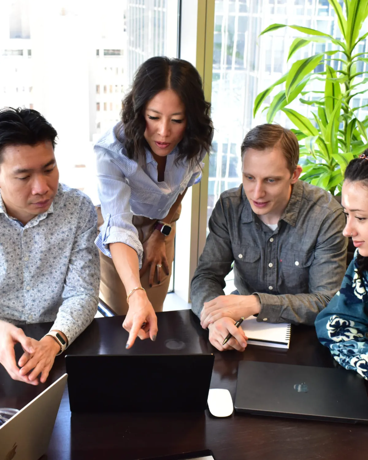 Denise leans over a boardroom table and points at a laptop screen while three colleagues follow along