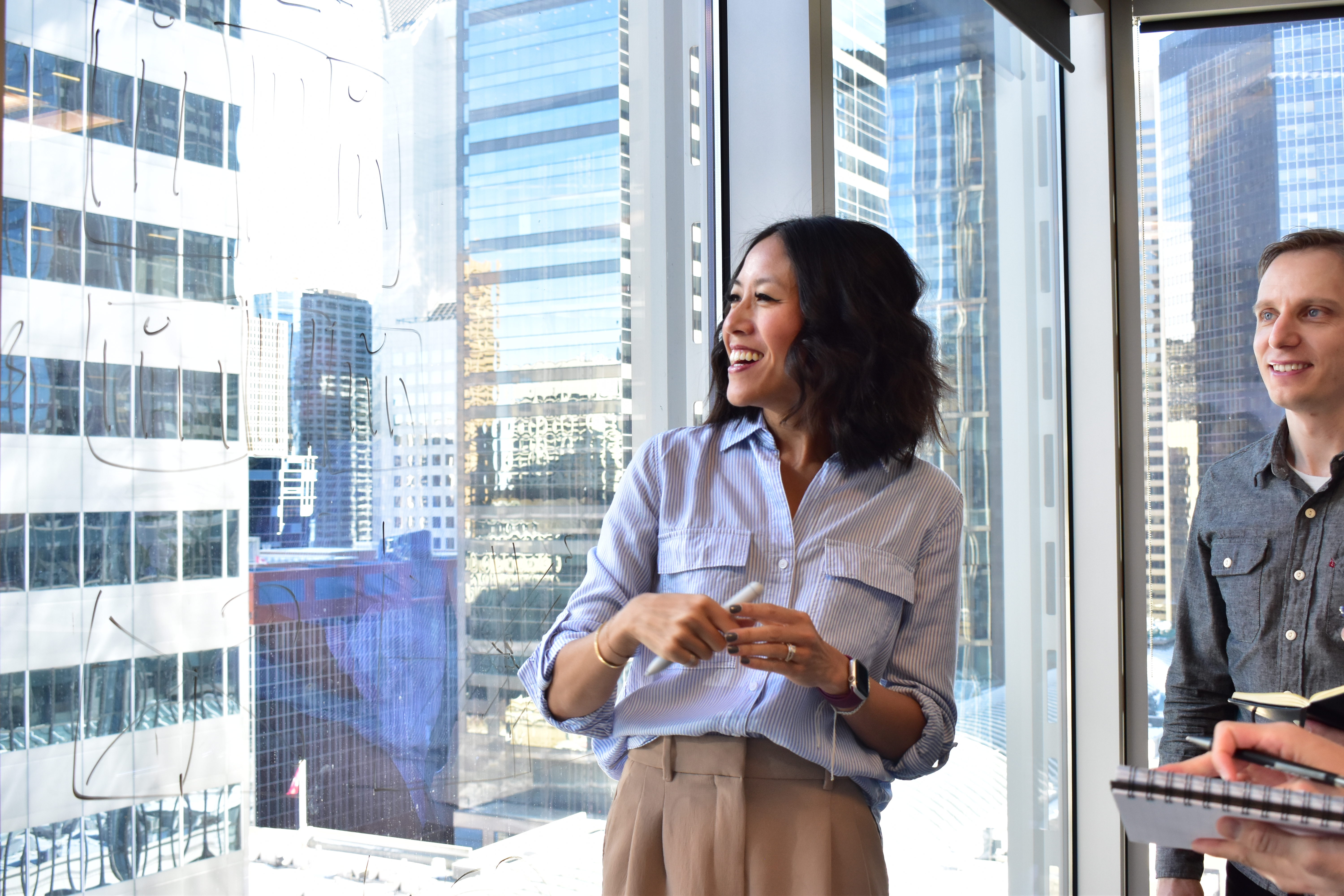 A woman in a light blue shirt smiles while holding a marker, standing near a large window overlooking a city skyline. A man in a gray shirt is also present, looking at her.