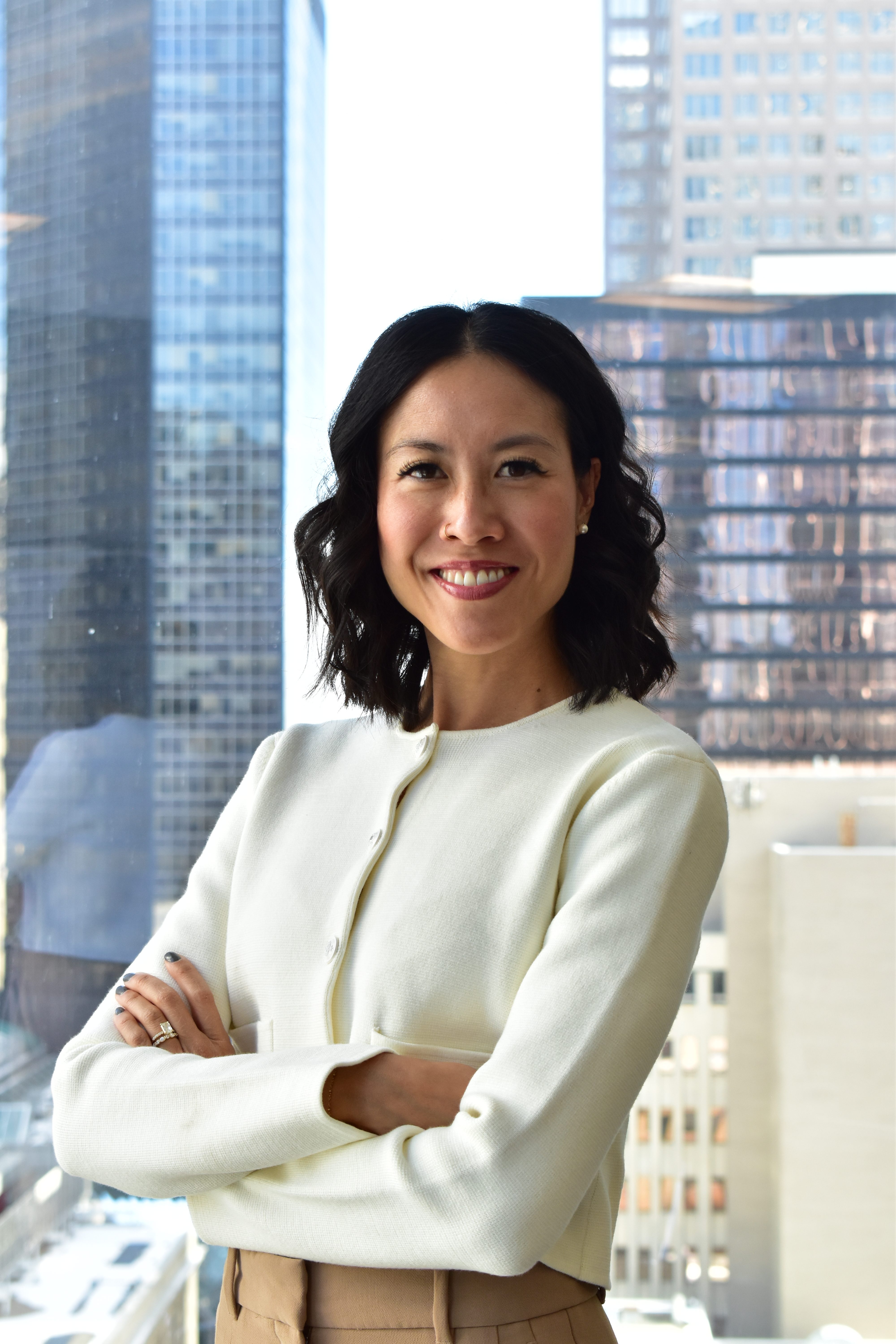 A woman smiling confidently with her arms crossed, wearing a cream-colored sweater and beige pants, standing in front of large windows with a cityscape background.